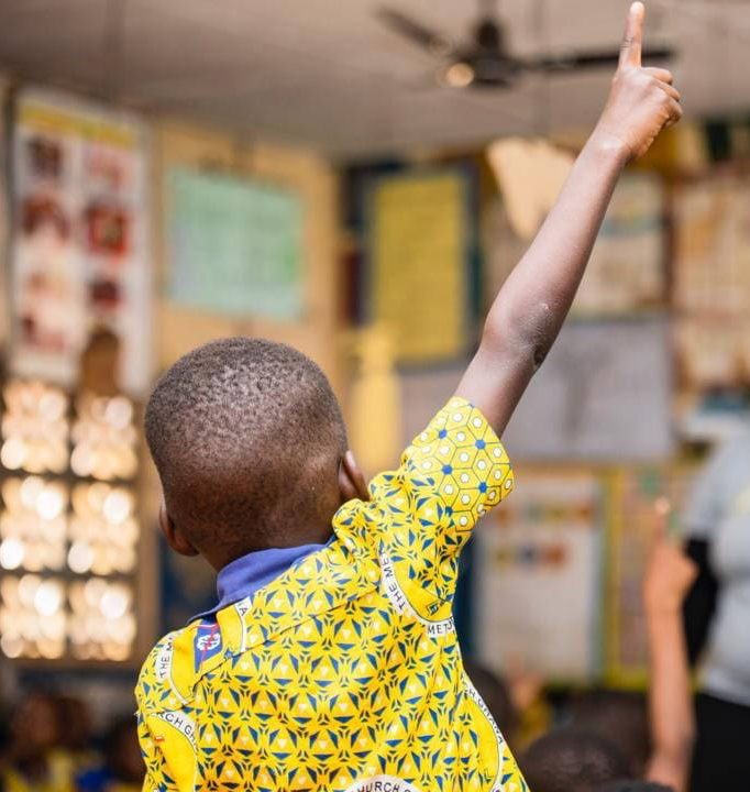 A young boy in a classroom with his back the camera. He has his hand pointing up and is wearing a yellow top.