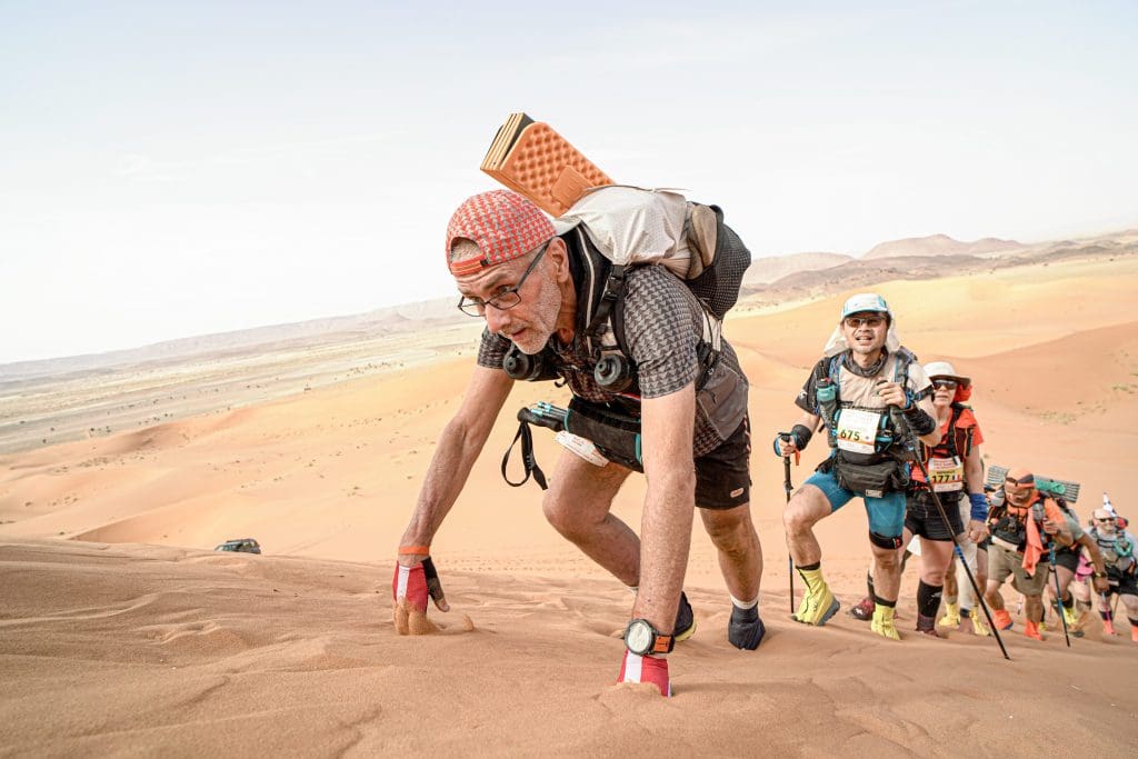 A man climbing up a steep dune in the desert. a line of people are behind him also climbing.