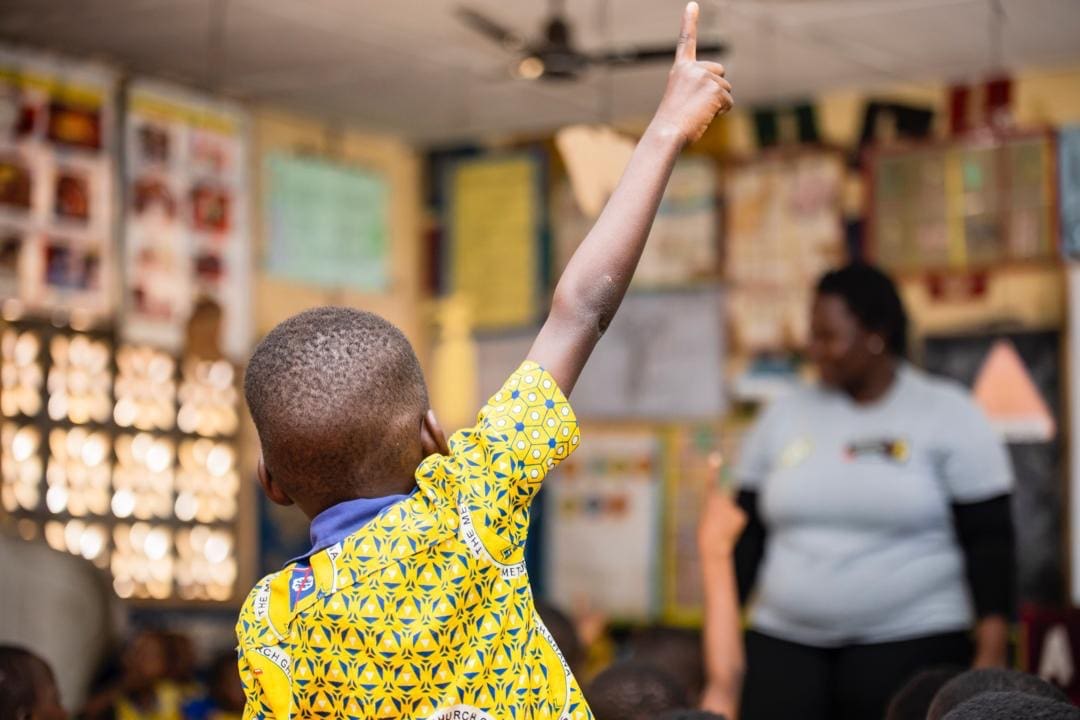 A young boy with his hand up in class, facing away from the camera. He wears a bright yellow top.